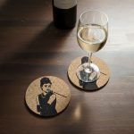 A cork coaster featuring Audrey Hepburn from Breakfast at Tiffany’s with a glass of white wine placed on top, alongside another matching coaster and a wine bottle on a wooden table.