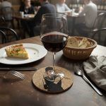 A cork coaster featuring Audrey Hepburn from Breakfast at Tiffany’s with a glass of red wine placed on top, set on a restaurant table with a slice of quiche, bread basket, and cutlery.