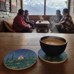 2 cork coasters featuring cottage and night sky design on a wooden table with a cup of tea kept on one of them, with people sitting indoors against a mountain view.
