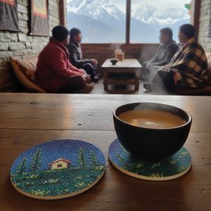 2 cork coasters featuring cottage and night sky design on a wooden table with a cup of tea kept on one of them, with people sitting indoors against a mountain view.