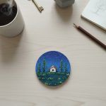 A cork coaster featuring a cottage and night sky design on a light wooden desk with a cup of coffee, pencil, notebook, and key.