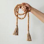 A hand holding a handcrafted wooden bead garland with jute tassels in natural finish, farmhouse decor.