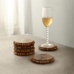 6 stacked round wooden bead coasters in natural finish beside champagne glass on table.