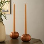 Rose gold metallic pumpkin-shaped taper candle holders with beige candles on wooden table beside vase with greenery.
