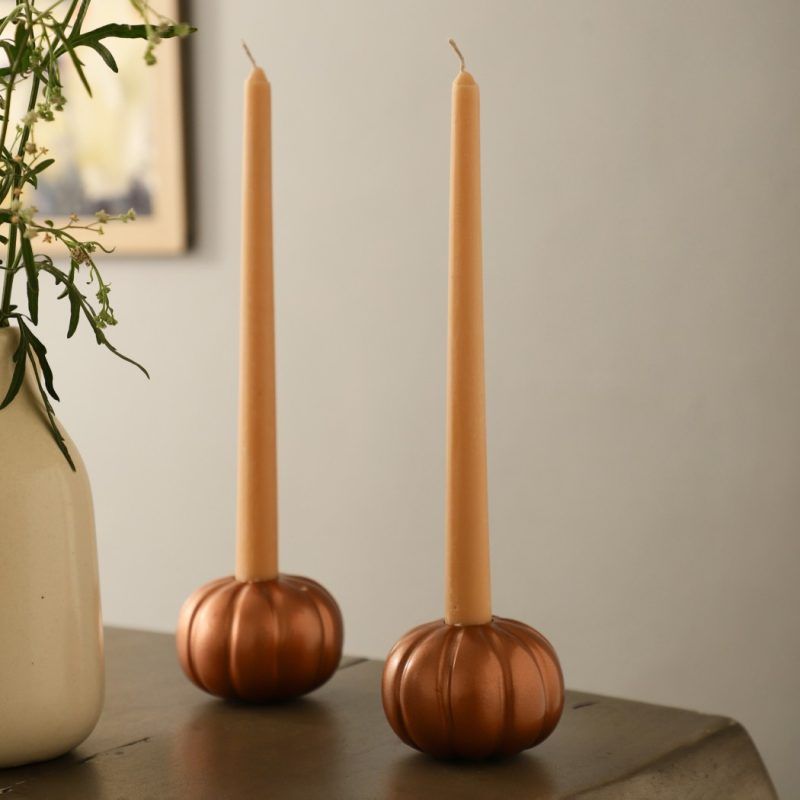 Rose gold metallic pumpkin-shaped taper candle holders with beige candles on wooden table beside vase with greenery.
