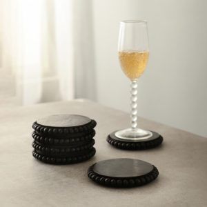 6 stacked round wooden bead coasters in black finish with champagne glass on table.