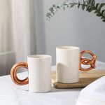 Two ceramic mugs with unique handles: one like churros and one pretzel-shaped in brown glaze, on white table with eucalyptus overhead.