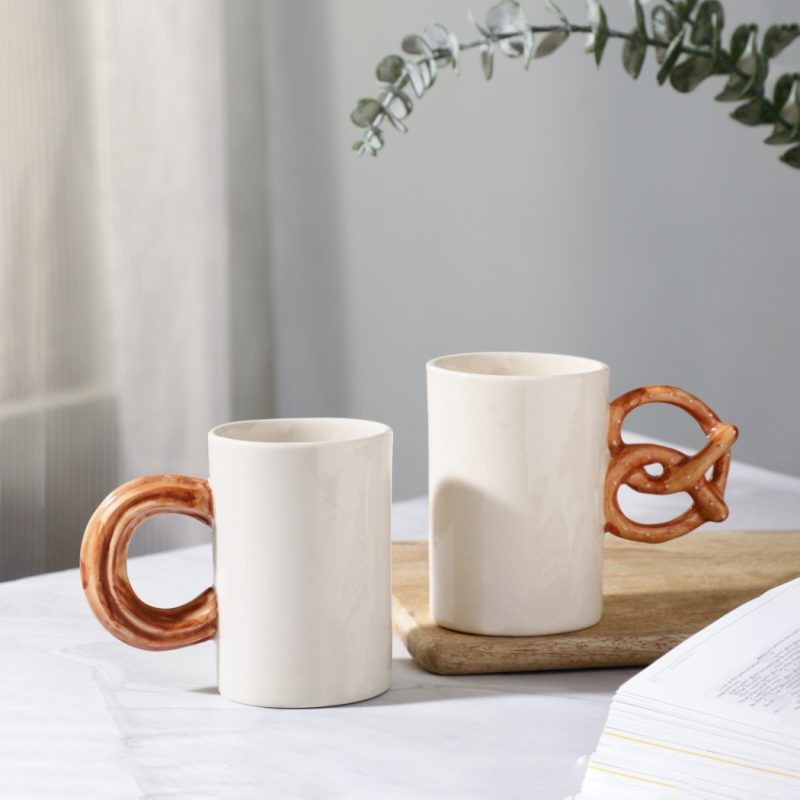 Two ceramic mugs with unique handles: one like churros and one pretzel-shaped in brown glaze, on white table with eucalyptus overhead.
