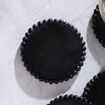 Close-up of a single black wooden coaster with beaded edge on white marble surface, three others partially visible.