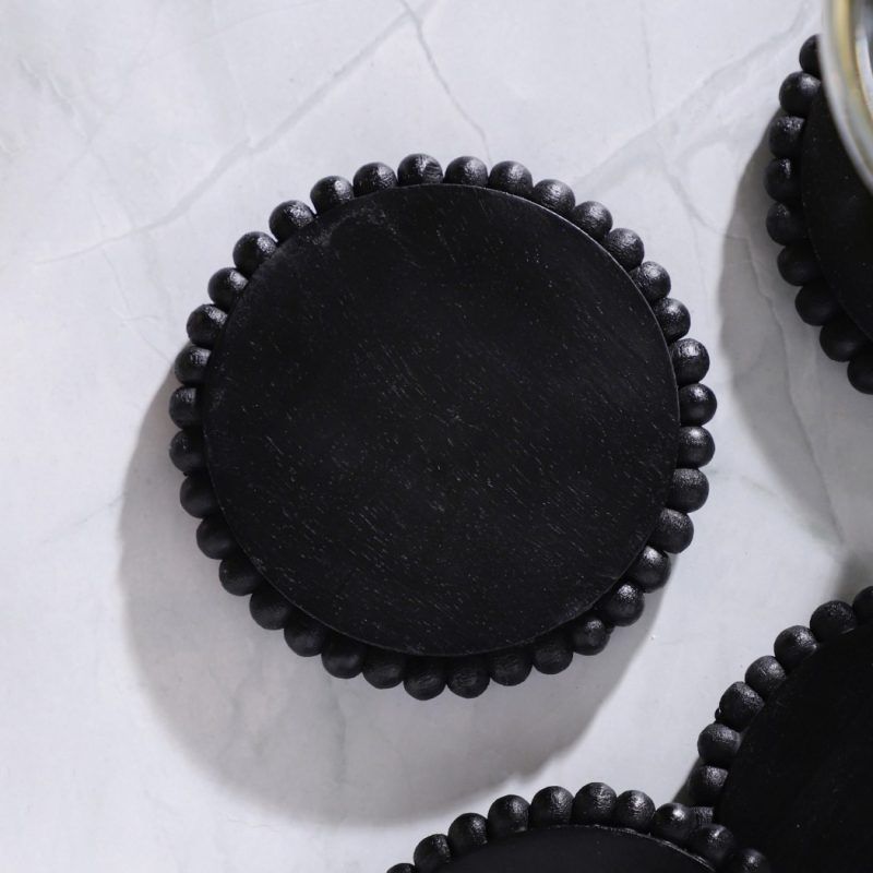 Close-up of a single black wooden coaster with beaded edge on white marble surface, three others partially visible.