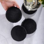 Four black wooden coasters with beaded edges, one held by hand, another supporting a glass of white wine with lemon slice on marble surface.