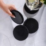 Four black wooden coasters with beaded edges, one held by hand lifting one to show underside, another supporting a glass of white wine with lemon slice on marble surface.