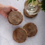 Set of four natural wooden beaded edge coasters on marble table, one held by hand, wine glass on another.