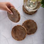 Set of four natural wooden beaded edge coasters on marble table, one held by hand, wine glass on another.