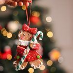 Hand holding the female figure from the Christmas tree gingerbread ornaments set of 2, featuring a red dress, red bow, and green candy cane against a bokeh light background.