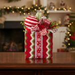 Front view of a fabric-wrapped holiday gift box decoration featuring a "Ho Ho Ho" ribbon and frosted greenery.