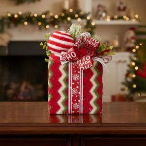 Front view of a fabric-wrapped holiday gift box decoration featuring a "Ho Ho Ho" ribbon and frosted greenery.