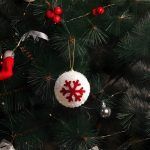 Close-up of a white textured frosted snowball ornament with a red snowflake design hanging on a green Christmas tree branch next to red berries.