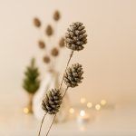 Close-up detail of glittered sequin pine cone sticks showing the textured metallic finish, with blurred warm holiday lights and a small tree in the background.