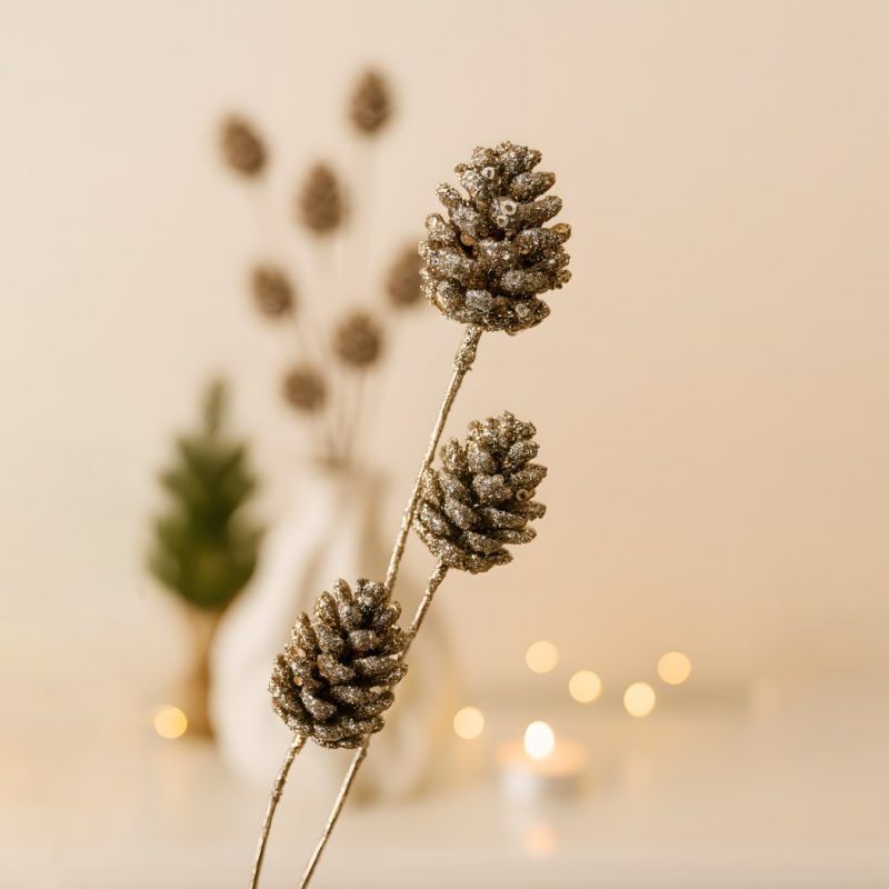 glittered-sequin-pine-cone-sticks-close-up-detail Close-up detail of glittered sequin pine cone sticks showing the textured metallic finish, with blurred warm holiday lights and a small tree in the background.
