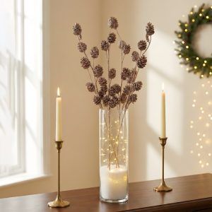 Elegant holiday centerpiece featuring glittered sequin pine cone sticks in a tall glass vase filled with fairy lights and faux snow, flanked by gold candlesticks.