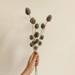 A hand holding a set of glittered sequin pine cone sticks against a beige background, demonstrating the stem length and realistic shape.