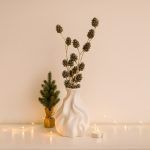 Set of glittered sequin pine cone sticks arranged in a modern white sculptural vase, styled on a table with a miniature Christmas tree and tea light candle.