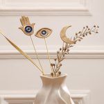 Close-up of the Golden Persian charm set (Moon, Eye, Hamsa) displayed in a modern white vase with dried botanicals.