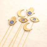 Flat lay showing the complete set of 6 golden metal sticks: two Crescent Moons, two Hamsa Hands, and two Evil Eyes with blue enamel details.