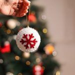A hand holding a white glittery snowball ornament with a red snowflake center, displaying the size against a blurred background of Christmas tree lights.