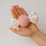 A hand holding a pastel pink candy Christmas ornament to show size and scale. The pink center has white piped lines, and the ends are covered in multicolored faux sprinkles.