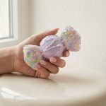 Close-up of a hand holding a sparkling lavender candy-shaped Christmas ornament. The ornament features a frosted sugar texture, white icing details, and pastel sprinkles on the ends.
