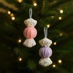 A pink and a violet toffee-shaped ornament hanging on a Christmas tree with glowing lights in the background. The pastel ornaments contrast against dark green pine branches for a whimsical holiday look.