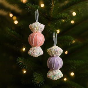 A pink and a violet toffee-shaped ornament hanging on a Christmas tree with glowing lights in the background. The pastel ornaments contrast against dark green pine branches for a whimsical holiday look.