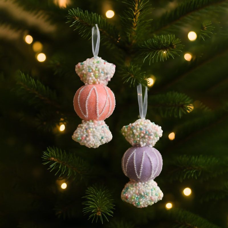A pink and a violet toffee-shaped ornament hanging on a Christmas tree with glowing lights in the background. The pastel ornaments contrast against dark green pine branches for a whimsical holiday look.