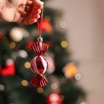 Close up detail of a red candy shaped Christmas hanging showing white glitter stripes held against a blurred Christmas tree background.