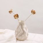 A set of 3 golden lotus blossom metal sticks displaying a minimalist look in a white ceramic vase, isolated on a clean background.