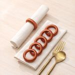 Flat lay display showing a set of burnt orange yarn napkin rings arranged on folded linen napkins with gold cutlery.