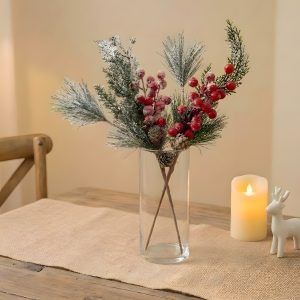 A dining table setting featuring a glass vase with frosted pine and berry stems, surrounded by a lit candle and a white reindeer figurine.