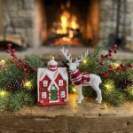 A festive Christmas ornament set featuring a red snowy cottage and a white reindeer with a red scarf, displayed on a wooden table with pine branches and a cozy fireplace in the background.