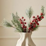 A close-up detail shot of frosted red berries, pine needles, and small pinecones arranged in a white wavy vase, highlighting the realistic snowy texture of the stems.