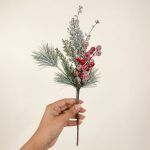 A hand holding a single stem of frosted pine needles and red berries to show the realistic texture and detail of the vase filler.