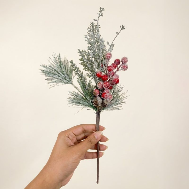 A hand holding a single stem of frosted pine needles and red berries to show the realistic texture and detail of the vase filler.