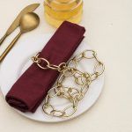 Stack of four champagne gold chain link style napkin rings on a dining table with glass decor props.