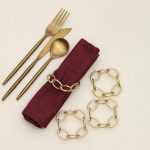 Flat lay of Glimpse Homes champagne gold metal knot napkin ring with gold cutlery and burgundy linen napkin.