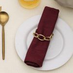 Elegant dining plate setting featuring a Glimpse Homes champagne gold metal knot napkin ring on a maroon napkin.