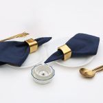 A pair of Glimpse Homes Golden Cuff Napkin Rings styled with navy napkins and gold flatware for a formal dinner setup.