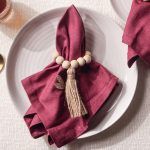 Top-down view of the Glimpse Homes Wooden Bead Napkin Ring on a burgundy napkin, highlighting the natural wood grain and tassel.