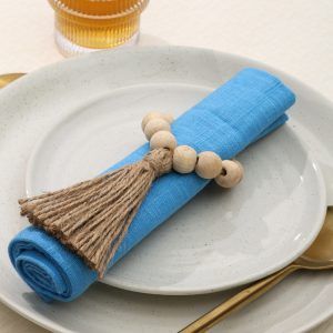 Close-up top view of a Glimpse Homes Wooden Bead Napkin Ring with a jute tassel, styled on a blue napkin with gold cutlery.