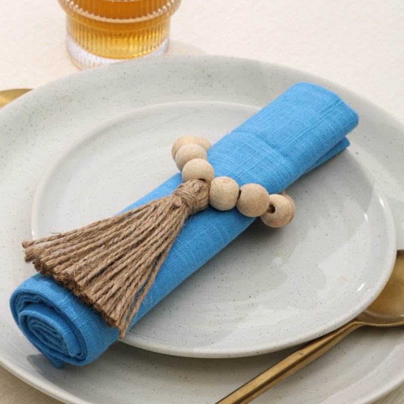 Close-up top view of a Glimpse Homes Wooden Bead Napkin Ring with a jute tassel, styled on a blue napkin with gold cutlery.